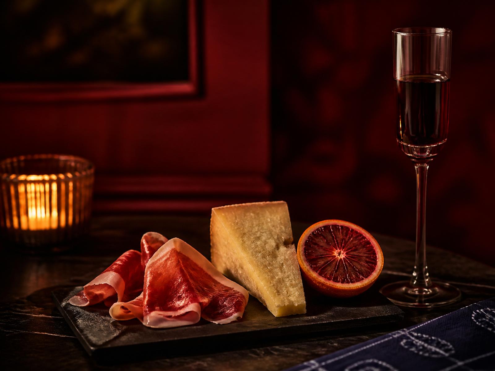 A small board of aged ham, aged cheese, and blood orange beside a slender flute in a candlelit red room.