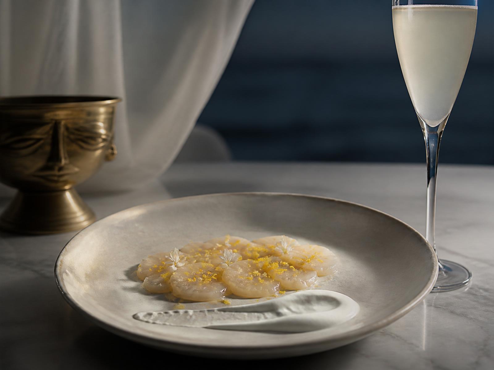 A single delicate scallop crudo plate on pale marble in moonlight, beside a slender flute.